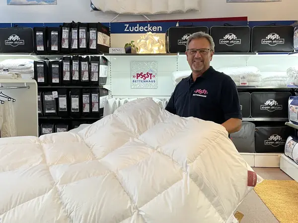 Mann präsentiert Daunendecke im Bettenfachgeschäft vor Regal mit verschiedenen Zudecken.